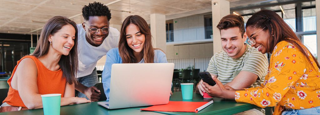 A group of happy young people looking at a phone and computer. Refer a friend to Blue hero image.