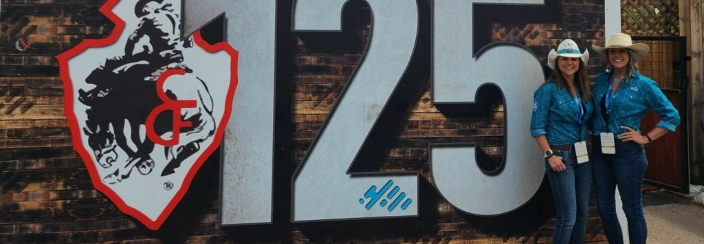 Cartel por los 125 años de Cheyenne Frontier Days y Blue Federal Credit Union con 2 empleados de Blue de pie junto a él.