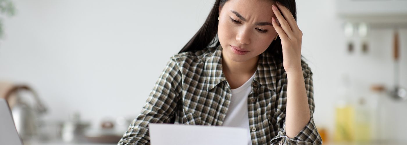 A woman in a white t-shirt and dark plaid overshirt examines a piece of mail with a furrowed brow and her hand on her head.