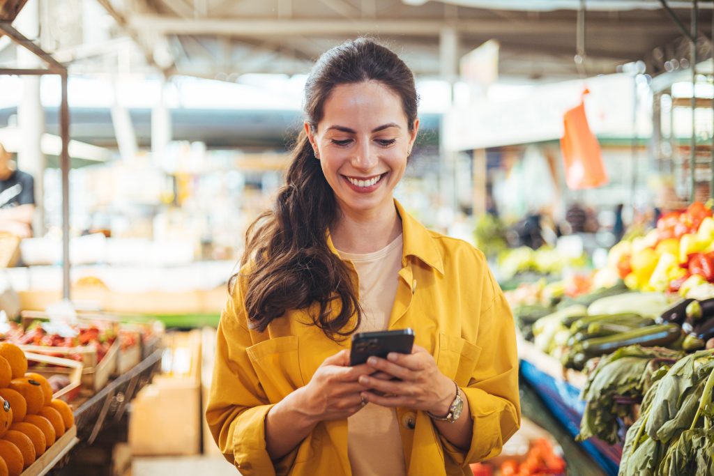 Mujer en el mercado de agricultores comprando, enviando un mensaje de texto por su smartphone. Joven alegre en el mercado. La chica sonriente decidió cocinar una comida deliciosa y saludable